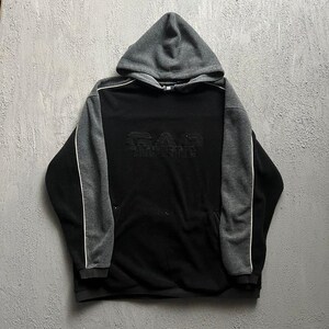 May include: A black and charcoal gray hooded sweatshirt featuring long sleeves with white piping accents. The front displays the text GAP ATHLETIC embroidered in a tonal black thread across the chest. The garment includes a hood and ribbed cuffs.