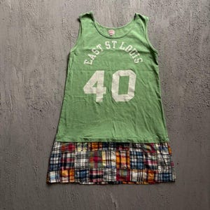 May include: A sleeveless green tank top dress featuring the white text EAST ST LOUIS arched over the large white number 40. The lower portion of the garment consists of a multi-coloured plaid patchwork skirt with various squares of blue, red, and yellow.