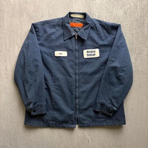 May include: A navy blue work jacket featuring a full-length front zip, a pointed collar, and long sleeves with buttoned cuffs. Two rectangular patches are sewn on the chest, one reading TIM and the other reading BODY SHOP in dark blue text.