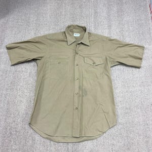 Könnte beinhalten: Ein kurzärmeliges, knopfleisten Hemd in Khaki. Das Hemd hat zwei Brusttaschen und einen Kragen. Der Stoff scheint ein strapazierfähiges Material im Arbeitsstil zu sein. Das Hemd liegt flach auf einer strukturierten Oberfläche.