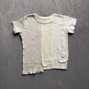 Puede incluir: Una camiseta de manga corta construida con tres paneles verticales en varios tonos de blanco roto y beige. El panel izquierdo presenta una textura de punto gofrado, mientras que los paneles central y derecho son de algodón liso. Texto impreso.