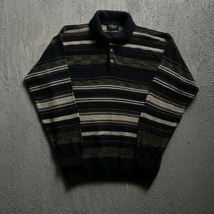 May include: A long-sleeve knit polo shirt featuring a horizontal striped pattern in shades of black, charcoal gray, and cream. The garment has a solid black collar, a two-button placket with silver-toned buttons, and ribbed cuffs and hem.