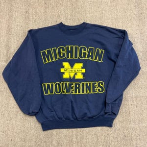 May include: A navy blue crewneck sweatshirt featuring the text MICHIGAN in yellow arched lettering above a large yellow block M logo with the word MICHIGAN centered inside it, followed by the word WOLVERINES in yellow capital letters.
