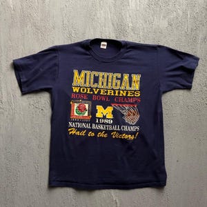 May include: A navy blue short-sleeve t-shirt featuring yellow and white text that reads Michigan Wolverines Rose Bowl Champs, National Basketball Champs, and Hail to the Victors, with a 1989 Rose Bowl logo, a yellow M, and a basketball hoop graphic.