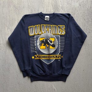 Könnte beinhalten: Ein langärmeliges, marineblaues Sweatshirt mit dem Schriftzug WOLVERINES in Weiß und Gelb, einem zentralen Wappen mit einem gelben M und einer schwarzen Vielfraß-Silhouette sowie dem Wort MICHIGAN in Gelb mit zwei Sternen auf der Vorderseite.