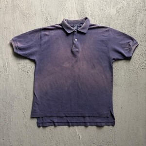 May include: A short-sleeved polo shirt in a faded, muted purple hue. The shirt features a classic collar, a two-button placket, and a small Gap brand label at the neckline. The hem is slightly longer in the back, creating a subtle drop-tail design.