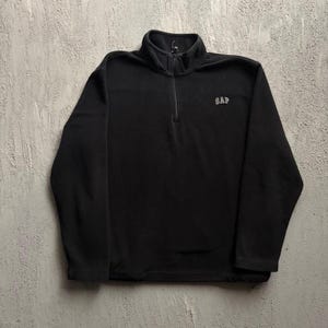 Pode incluir: Um suéter de fleece preto com gola alta, mangas compridas e um fecho de correr de um quarto de comprimento na parte central frontal. A palavra GAP está bordada em letras pequenas de cor clara na parte superior esquerda do peito.