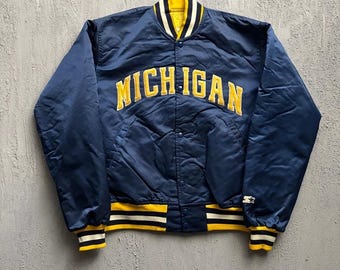 May include: A navy blue satin varsity jacket featuring the word MICHIGAN in yellow block letters across the chest. The jacket has a snap-button front, a ribbed collar, cuffs, and hem with yellow and white stripes, and a small Starter logo on the sleeve.