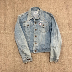 May include: A light blue denim jacket with a pointed collar, long sleeves, and a button-up front. The jacket features two chest pockets with buttoned flaps, copper-coloured buttons, and a Wrangler brand label on the inside neck area.