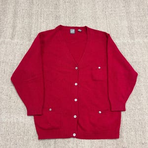 Könnte beinhalten: Roter Cardigan mit V-Ausschnitt, Knopfverschluss und zwei Vordertaschen. Der Pullover hat lange Ärmel und gerippte Bündchen und Saum. Das Gap-Logo ist auf der Rückseite des Kragens sichtbar.