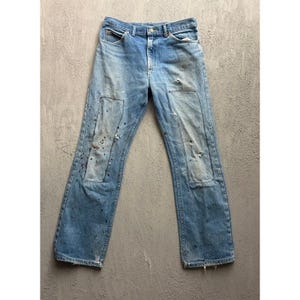 May include: Light blue denim jeans featuring a classic five-pocket design with a button fly closure. The fabric displays a distressed finish with rectangular patches on both knees, accompanied by scattered paint splatters in various dark colors.