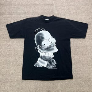 Op de afbeelding: Een zwart t-shirt met korte mouwen en een grote witte grafische print van het hoofd en de schouders van Homer Simpson. Het ontwerp maakt gebruik van een gestippelde schaduwtechniek voor diepte. Onderaan de afbeelding staat kleine tekst.