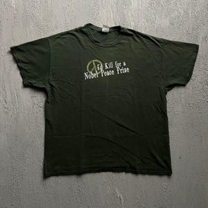 May include: A dark olive green short-sleeve t-shirt featuring a faded olive peace sign graphic and the white text "I'd Kill for a Nobel Peace Prize" printed on the chest. The shirt has a standard crew neckline and a relaxed, casual fit.