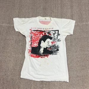 May include: White t-shirt with a graphic of Rick Springfield. The design features a black and white portrait within a red and black rectangular frame, with the text "RICK SPRINGFIELD" above. The shirt is vintage and shows signs of wear.