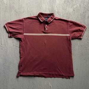Puede incluir: Un polo de manga corta en color granate con un cuello que presenta una raya blanca, una tapeta de tres botones y una raya horizontal blanca en el pecho. La prenda incluye una pequeña etiqueta de la marca Gap en el cuello y puños acanalados.
