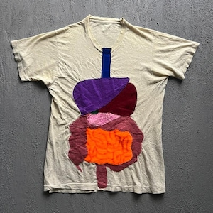 Könnte beinhalten: Ein cremefarbenes Kurzarm-T-Shirt mit einer großen, bunten Stoffapplikation auf der Vorderseite. Das Design zeigt ein stilisiertes menschliches Verdauungssystem mit einem blauen vertikalen Streifen sowie violetten, kastanienbraunen, rosafarbenen und leuchtend orangefarbenen Stoffteilen.