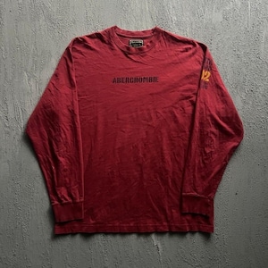 May include: A long-sleeve crew neck shirt in a deep red colour. The front features the text ABERCROMBIE printed in black across the chest. On the left sleeve, there is a graphic with the number 92 in yellow and additional black text.