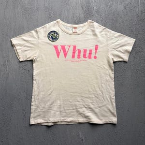 以下が含まれることがあります： 胸元に鮮やかなピンク色の「Whu!」という文字がプリントされた、クリーム色の半袖Tシャツ。クルーネック付近の左上部には、緑、青、白のディテールが施された小さな円形のワッペンが付いています。
