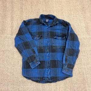 Puede incluir: Una camisa de manga larga con botones y un patrón de cuadros en azul y negro. La camisa incluye un cuello estándar, dos bolsillos en el pecho con solapas abotonadas y puños con botones. El nombre de la marca GAP aparece en la etiqueta interior.