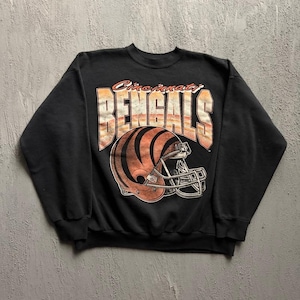 May include: A black crewneck sweatshirt featuring a graphic print of a football helmet with orange and black tiger stripes. Above the helmet, the text reads Cincinnati BENGALS in large, distressed orange and white block lettering.