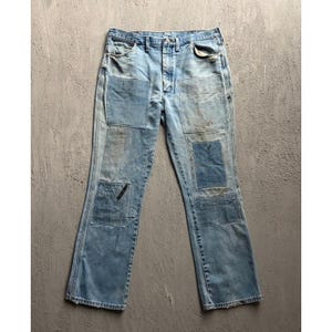 May include: Light blue denim jeans featuring a button fly closure, belt loops, and five pockets. The fabric displays a faded wash with multiple rectangular patches in varying shades of blue denim stitched onto the front of both legs.