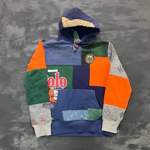 Puede incluir: Una sudadera con capucha y manga larga con un diseño de patchwork de paneles de tela azul marino, naranja, verde y gris. El frente muestra varios textos impresos que incluyen Polo Guide, Quality Dry Goods y Polo, con un parche bordado circular.