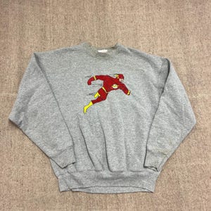 Może przedstawiać: Szara melanżowa bluza z okrągłym dekoltem, ozdobiona grafiką przedstawiającą postać komiksową The Flash w biegu. Postać ma na sobie czerwony kostium z żółtymi błyskawicami na klatce piersiowej, w talii oraz na butach.