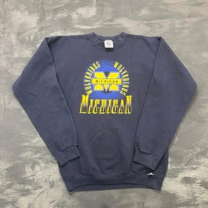 Könnte beinhalten: Ein marineblaues Sweatshirt mit Rundhalsausschnitt und einem Brustaufdruck, der ein gelbes M in Blockschrift, das Wort MICHIGAN in Gelb und Weiß sowie den Schriftzug WOLVERINES in einem kreisförmigen blau-gelben Farbverlauf zeigt.