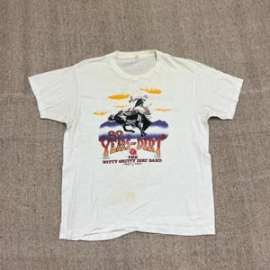 Puede incluir: Una camiseta blanca de manga corta con un gráfico de una persona montando un caballo encabritado. El texto dice 20 YEARS OF DIRT y THE NITTY GRITTY DIRT BAND. El diseño incluye nubes naranjas, montañas moradas y una pequeña rosa roja.