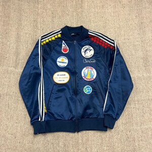 Op de afbeelding: Een marineblauw trainingsjack met rits, witte strepen op de mouwen, gele en rode sterren en diverse geborduurde patches, waaronder USA Swimming, Marlins Swim Club, Carino, Metro Swimmer en een kampioenschapsbadge uit 1983-84.