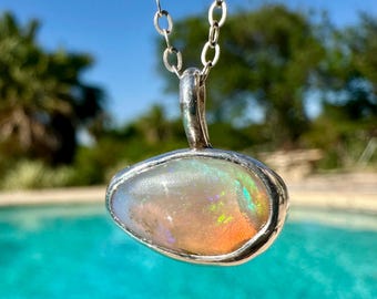 May include: A silver chain necklace with a pendant featuring a white opal gemstone set in a silver bezel. The opal has a rainbow of colors, including pink, green, and blue.