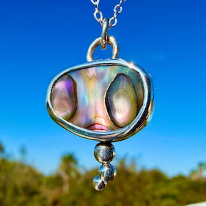 Peut inclure: Un collier pendentif en argent avec une incrustation de coquille d'ormeau ovale et irisée, sertie dans un cadre en argent poli. Le pendentif est suspendu à une chaîne en argent et comporte trois petites sphères en argent graduées.