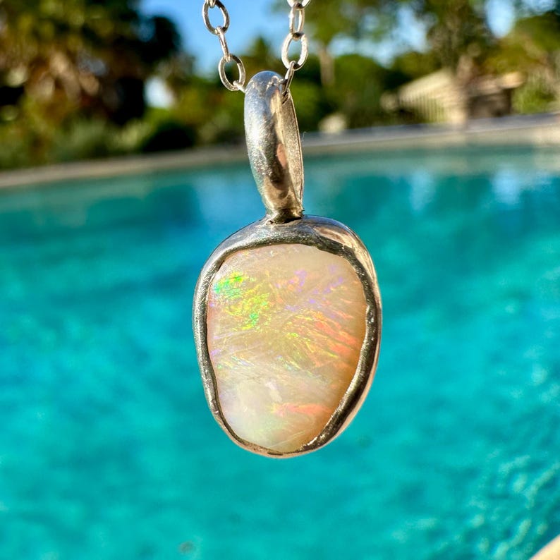 May include: A silver chain necklace with a pendant featuring a white opal stone set in a silver frame. The opal has a rainbow iridescent sheen.