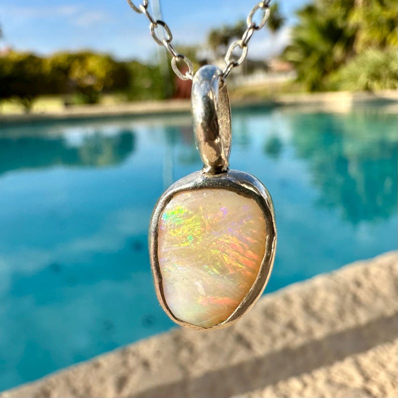 May include: A silver pendant necklace featuring a white opal gemstone. The opal displays iridescent rainbow colours.
