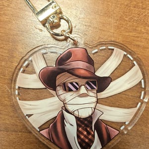 May include: A heart-shaped keyring with a cartoon illustration of a figure wrapped in bandages, wearing a hat and sunglasses. The figure is dressed in a suit and tie. The keyring has a gold-coloured clasp.