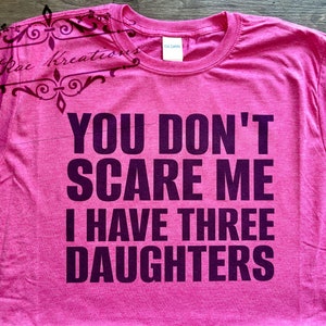 May include: A pink t-shirt with the text "YOU DON'T SCARE ME I HAVE THREE DAUGHTERS" in dark purple. The shirt has a crew neck and short sleeves. The brand name "GILDAN" is visible on the tag. The upper left corner has a decorative design with the text "K. Rae Kreations".