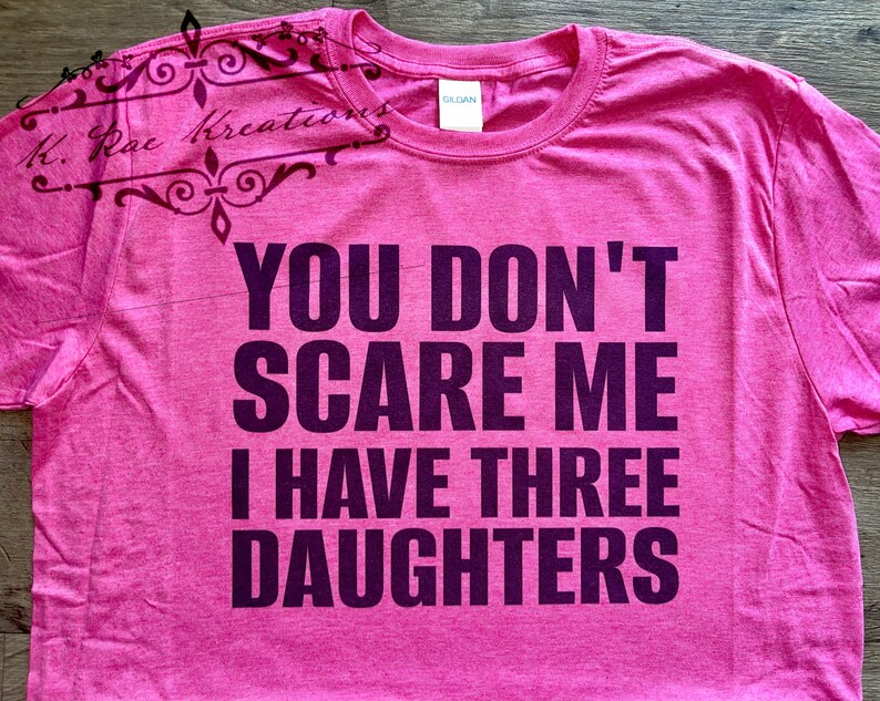 May include: A pink t-shirt with the text "YOU DON'T SCARE ME I HAVE THREE DAUGHTERS" in dark purple. The shirt has a crew neck and short sleeves. The brand name "GILDAN" is visible on the tag. The upper left corner has a decorative design with the text "K. Rae Kreations".