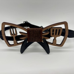 Pode incluir: Gravata borboleta de madeira com acabamento castanho, com desenhos recortados de um garfo e uma espátula. O centro tem uma textura de serapilheira, e a gravata borboleta é presa a uma faixa elástica preta. A gravata borboleta é suportada por duas peças pretas curvas.