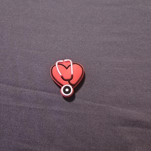 May include: A red heart-shaped charm with a white and red stethoscope design. The charm has a black outline and is set against a dark gray background. This medical-themed charm is likely for shoes or accessories.