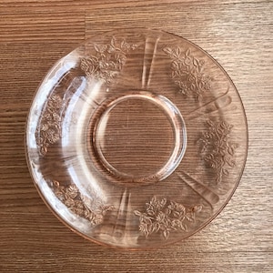 May include: A clear glass saucer with a delicate floral pattern. The saucer has a slightly pink hue and is perfect for serving tea or coffee.