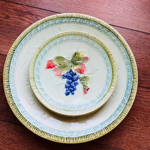 May include: Two ceramic plates, one nested inside the other, with a light green and yellow color scheme. The plates feature a raised grape design with blue grapes, green leaves, and red accents. The rims have a decorative border.