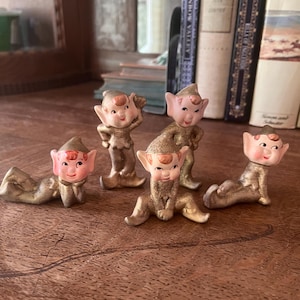 May include: A set of four small elf figurines with painted pink faces, pointed ears, and gold-toned bodies. Each elf is depicted in a different pose, including lying down, sitting, and standing, all featuring detailed facial expressions.