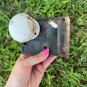 May include: Vintage door lock hardware with a white doorknob and a black metal plate. The lock shows signs of age and wear, with rust and discoloration. The hardware is held in a hand with pink nail polish.