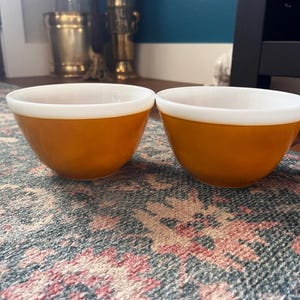 May include: Two vintage orange and white mixing bowls. The bowls have a rounded shape with a wide, white rim. The exterior is a solid, warm orange color. The bowls are sitting on a patterned rug.