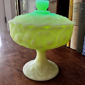 May include: A vintage, light green glass candy dish with a lid. The dish has a pedestal base and is decorated with a leaf pattern. The lid has a flower-shaped handle. The dish is sitting on a wooden surface.