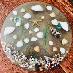 May include: A round, clear resin coaster with embedded seashells, a starfish, and small pebbles. The coaster features white and tan seashells, green seaweed, and a variety of colorful pebbles around the edge. The design evokes a coastal theme.