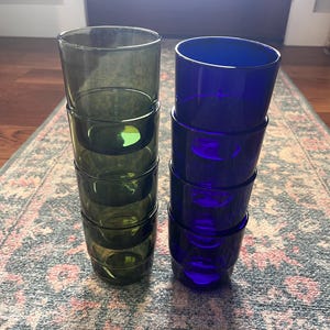 May include: Two stacks of drinking glasses. One stack is olive green, the other is a deep blue. Each stack has five glasses. The glasses are cylindrical with a slightly flared rim. The glasses are sitting on a patterned rug.
