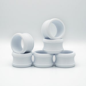 May include: A stack of six white ceramic rings, each with a smooth, rounded surface. The rings are arranged in a pyramid shape, with the largest ring at the bottom and the smallest ring at the top.