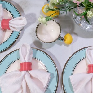 May include: Three place settings with white napkins and coral napkin rings on blue and white plates. The plates have a gold trim.