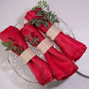 May include: Three red napkins with a sparkly texture are rolled up and secured with beige napkin rings. The napkins are arranged on a clear glass plate with a decorative pattern.